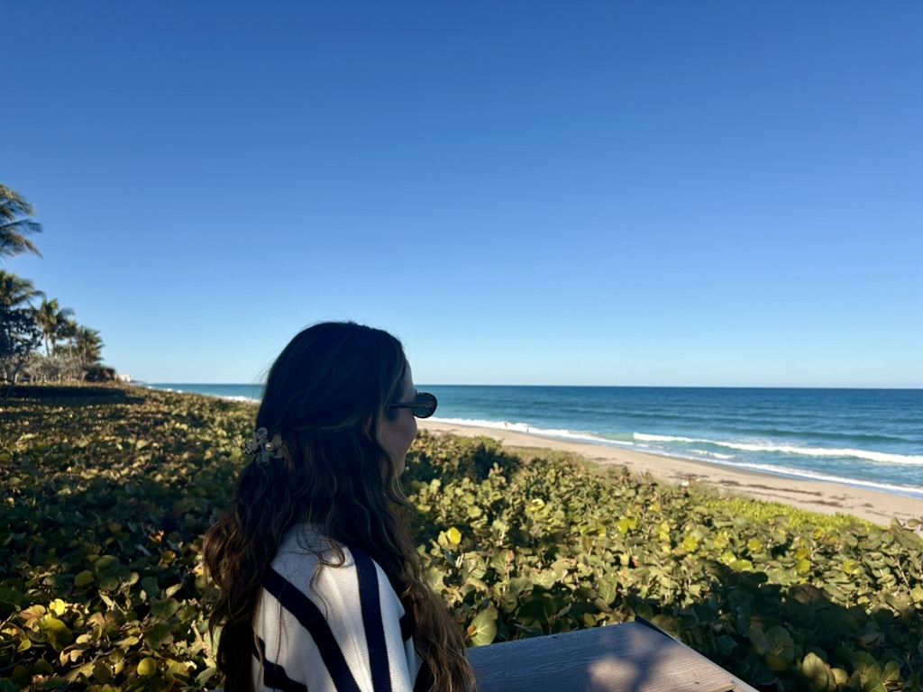 A woman with long hair wearing sunglasses, sitting by the beach, looking out at the ocean under a clear blue sky.